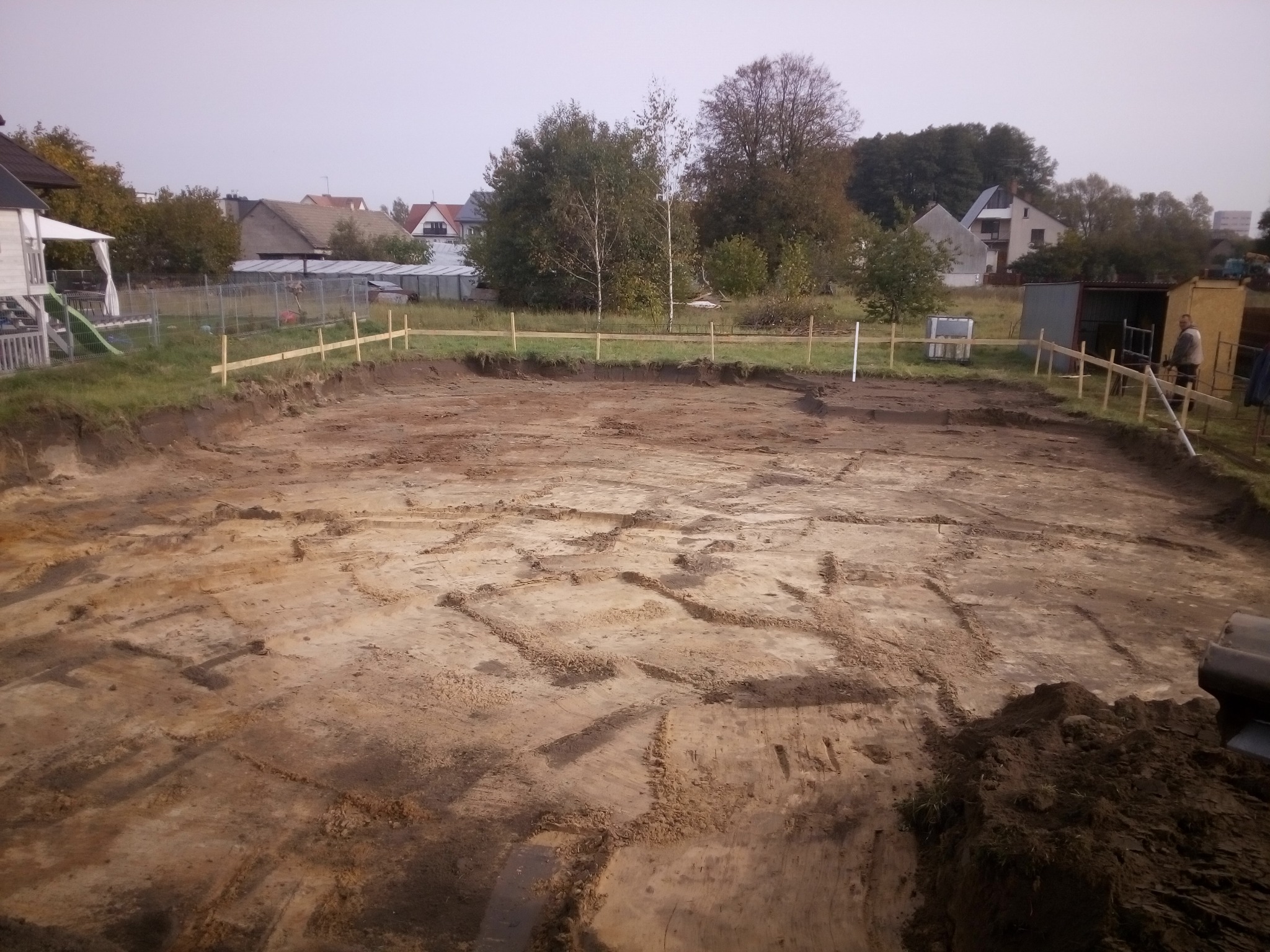 Wykop pod fundamenty domu jednorodzinnego, widoczne ślady po pracy koparki, drewniane paliki wyznaczające obrys fundamentów, w tle zabudowa mieszkalna.