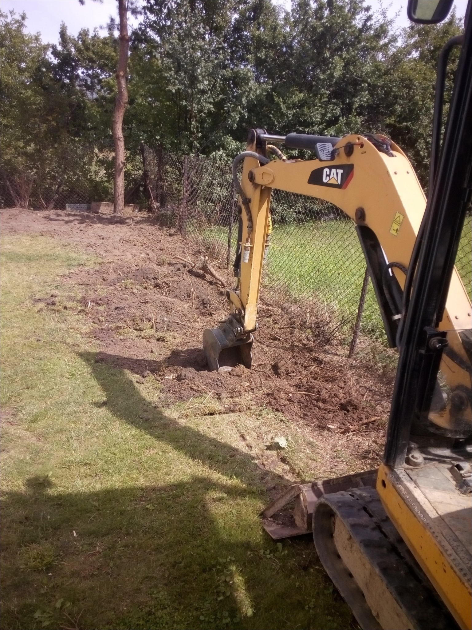Żółta mini koparka CAT podczas prac ziemnych na działce z siatką ogrodzeniową w tle.