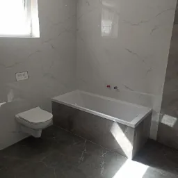 Nowoczesna łazienka z białymi marmurkowymi płytkami ściennymi, szarą podłogą z żyłami, białą wanną z szarym obramowaniem i podwieszaną toaletą.