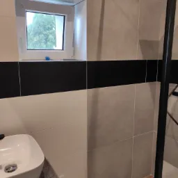 Nowoczesna łazienka z umywalką nablatową, kabiną prysznicową z czarną ramą i oknem z widokiem na drzewo, wykończona płytkami w odcieniach bieli, szarości i czerni.