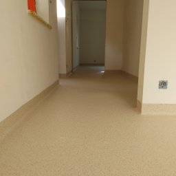 Floor Pro - Beżowa wykładzina PCV ułożona w korytarzu, z widocznymi listwami przypodłogowymi i gniazdkiem elektrycznym bez osprzętu. W tle widoczne kolejne pomieszczenia.