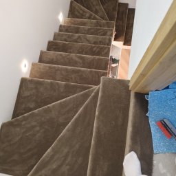 WOJCIECH MARKIEWICZ FLOOR PRO - Kaskadowe schody wyłożone beżową wykładziną, widziane z góry. Fragment stopy w skarpetce na dole kadru. Oświetlenie punktowe na ścianie. Widoczne narzędzia pracy.