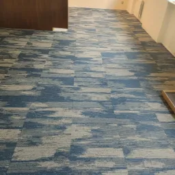 Wzorzysta, błękitno-szara wykładzina dywanowa w płytkach, ułożona w przestronnym wnętrzu z oknem i jasnymi ścianami. Widoczny fragment drzwi po lewej stronie.