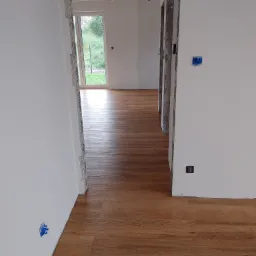 Ukończona podłoga z paneli w jasnym wnętrzu. Widoczne detale wykończenia i świeżo pomalowane ściany. Przejście do kolejnego pomieszczenia z widokiem na zieleń.