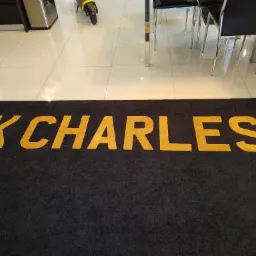 Czarna wykładzina z napisem 'K CHARLES' w złotym kolorze, leżąca na jasnych płytkach. W tle fragment skutera i krzesła.