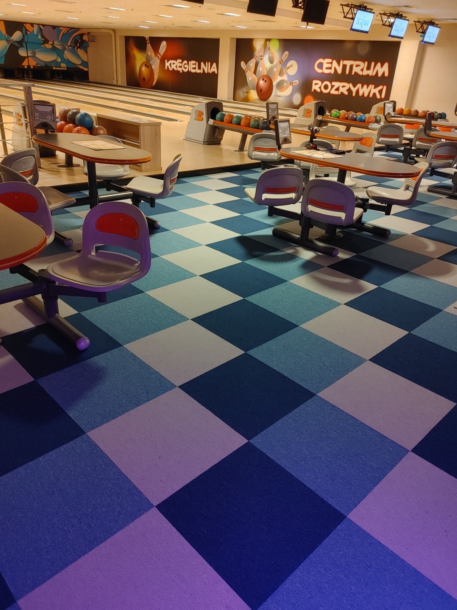 Wnętrze kręgielni z widocznymi torami, kulami do gry, stolikami z krzesłami i podłogą w niebiesko-białą szachownicę.