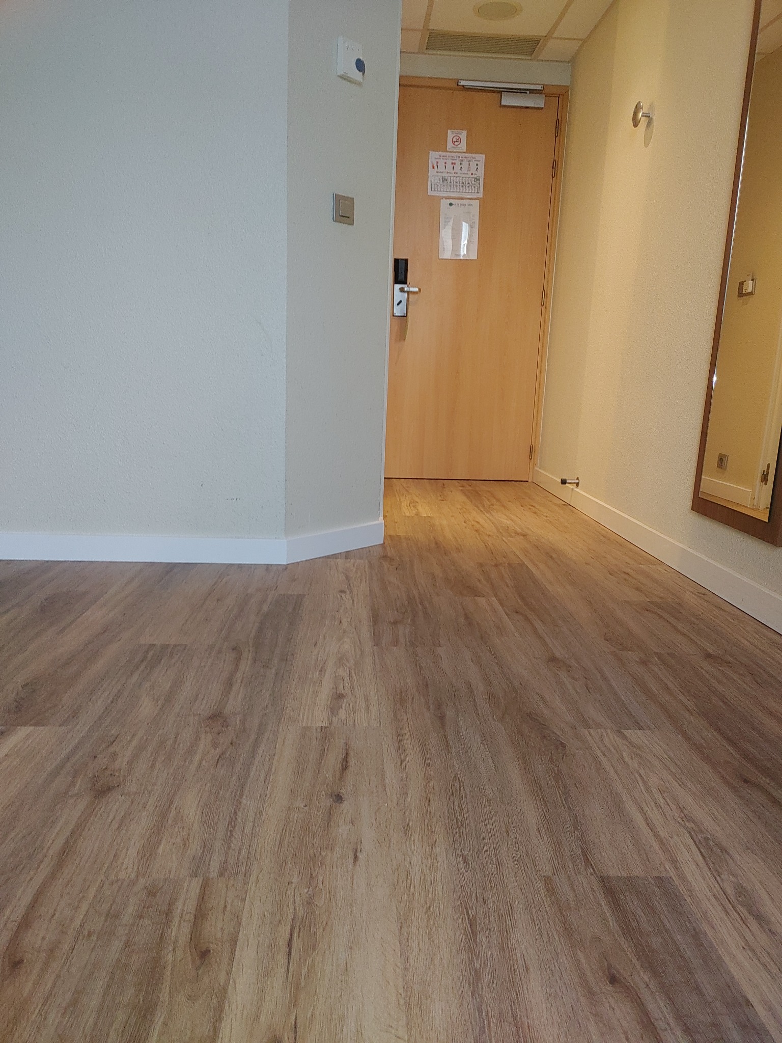 Jasnobrązowa podłoga z paneli imitujących drewno, widoczna przy ścianie z białymi listwami przypodłogowymi i drzwiami wejściowymi do pokoju hotelowego.