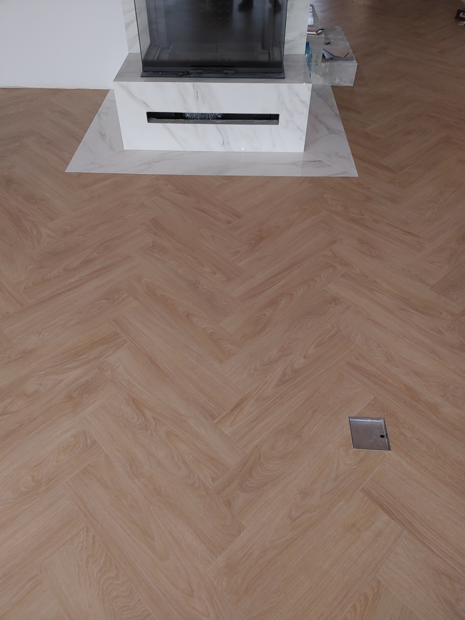 Jasne panele ułożone we wzór jodełki francuskiej, widoczna kratka wentylacyjna w podłodze oraz fragment kominka z marmurowym wykończeniem.