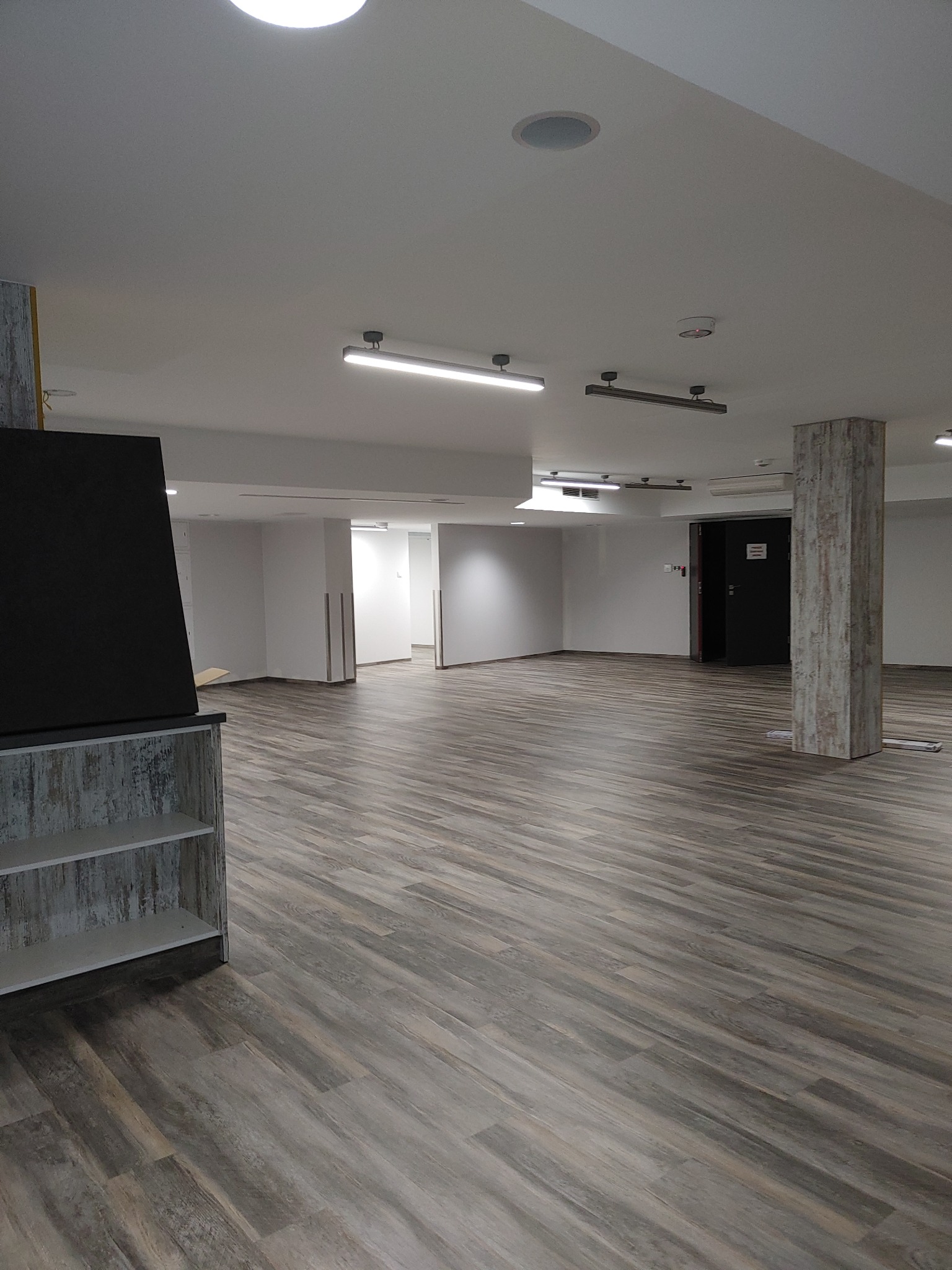 Duża przestrzeń z szarą podłogą imitującą drewno, oświetlona lampami sufitowymi, widok na dalsze pomieszczenia przez otwarte przejścia.