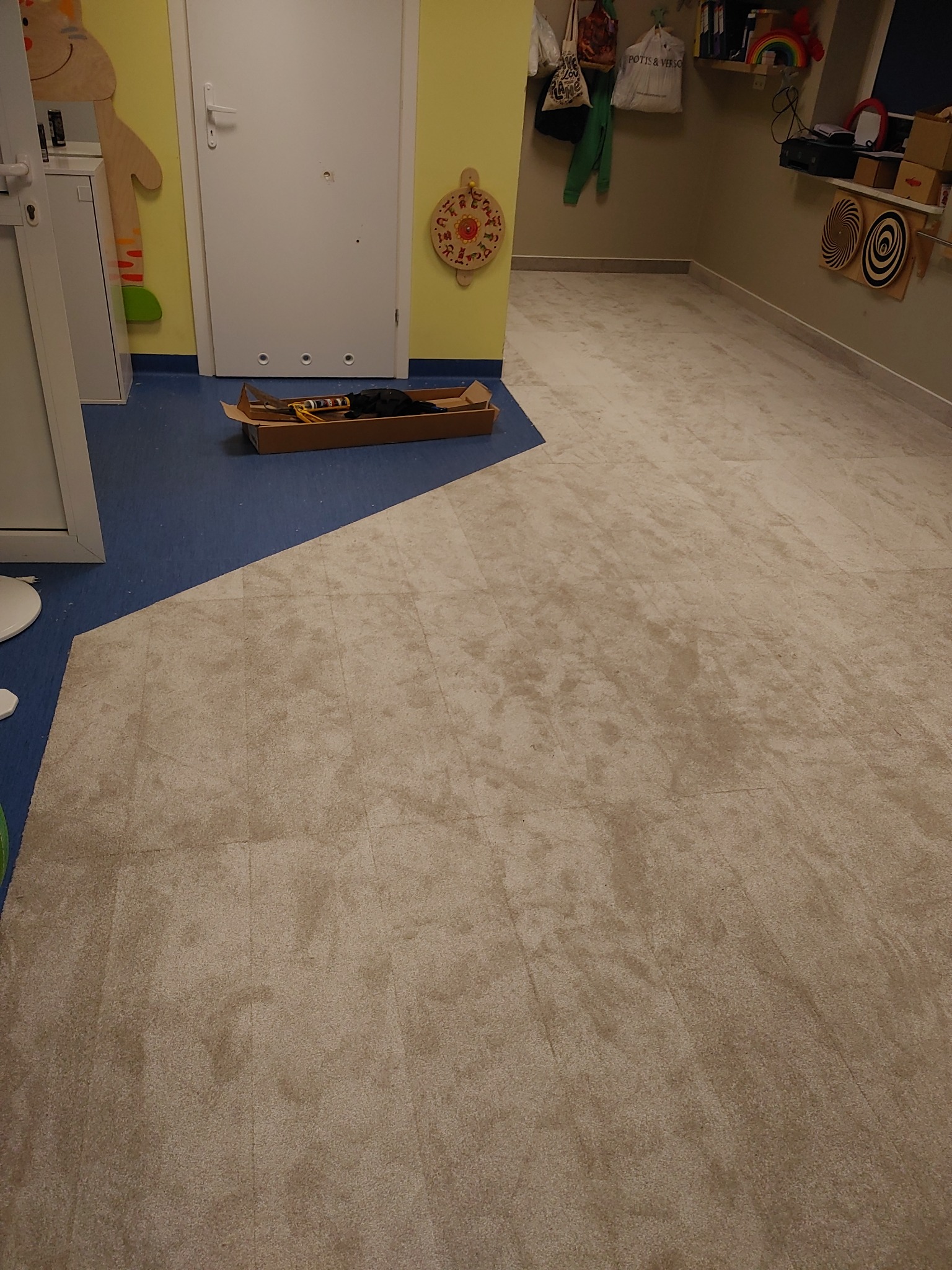 Beżowa podłoga wyłożona płytkami imitującymi wykładzinę dywanową w pokoju dziecięcym, z narzędziami w kartonie przy białych drzwiach i niebieską podłogą w tle.
