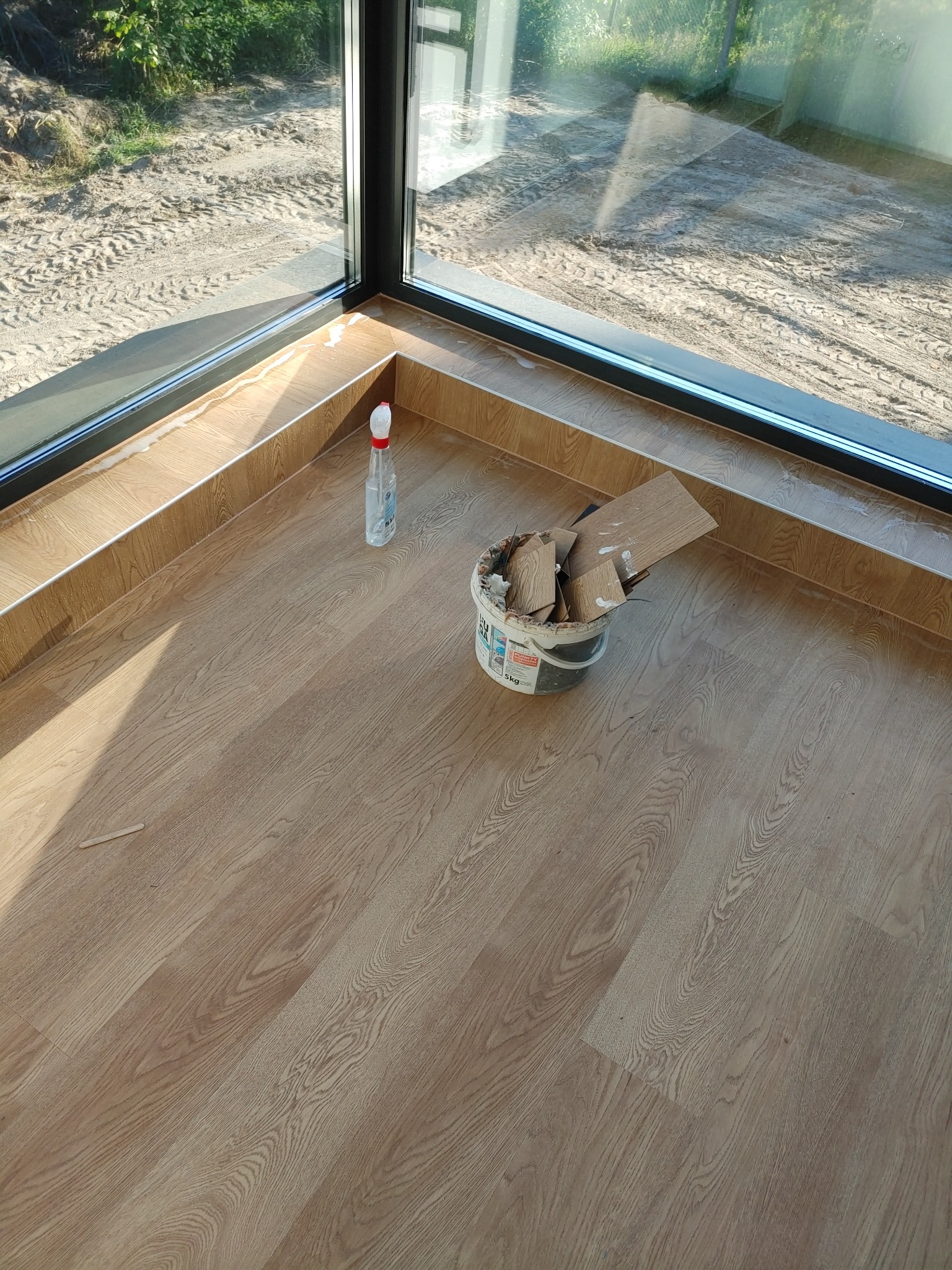 Ułożone panele podłogowe w rogu pokoju z dużym oknem, widoczny kubeł z resztkami paneli i butelka płynu do czyszczenia