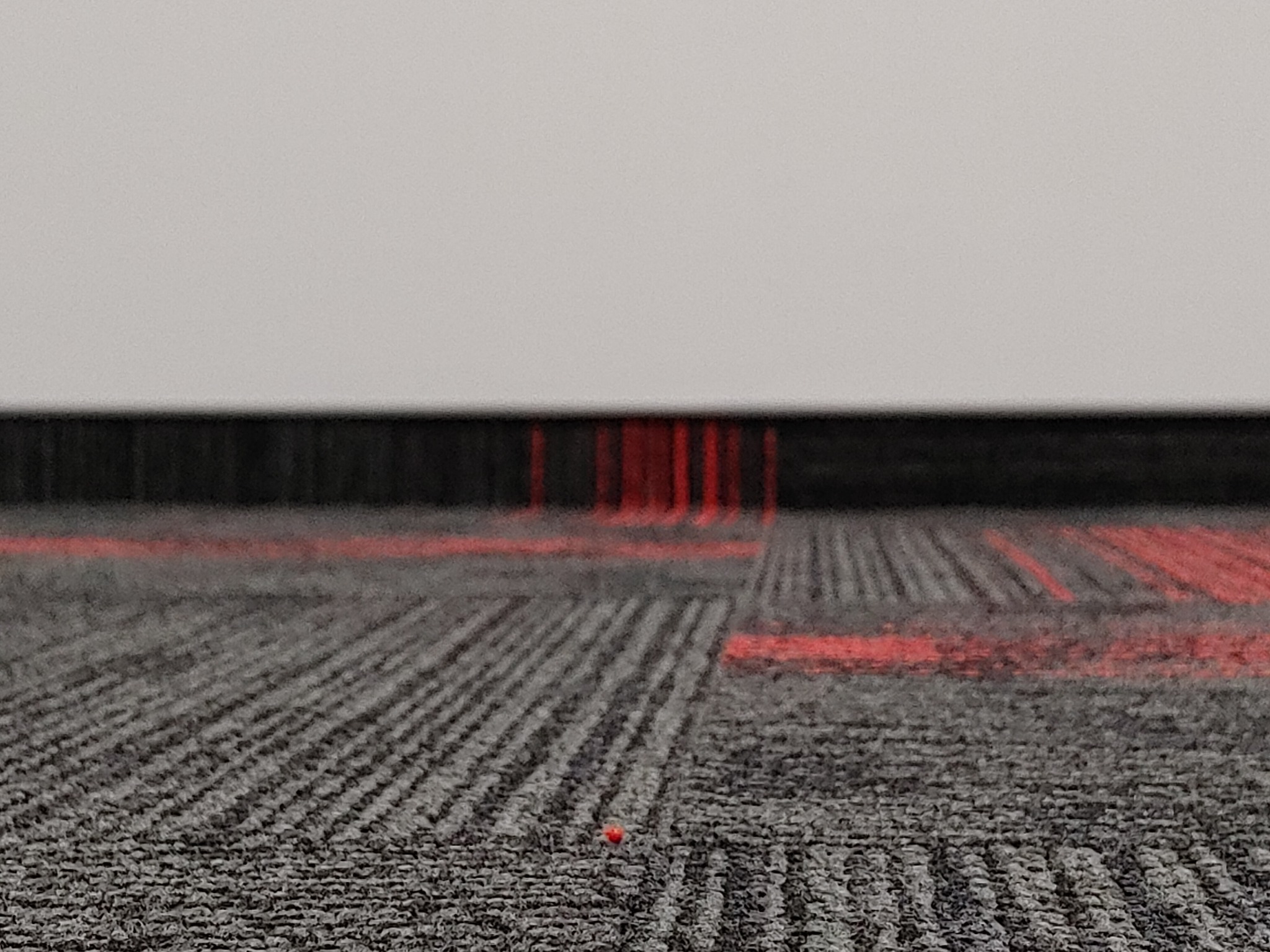 Zbliżenie na szarą wykładzinę dywanową z czerwonymi akcentami, widoczny fragment ściany u góry.