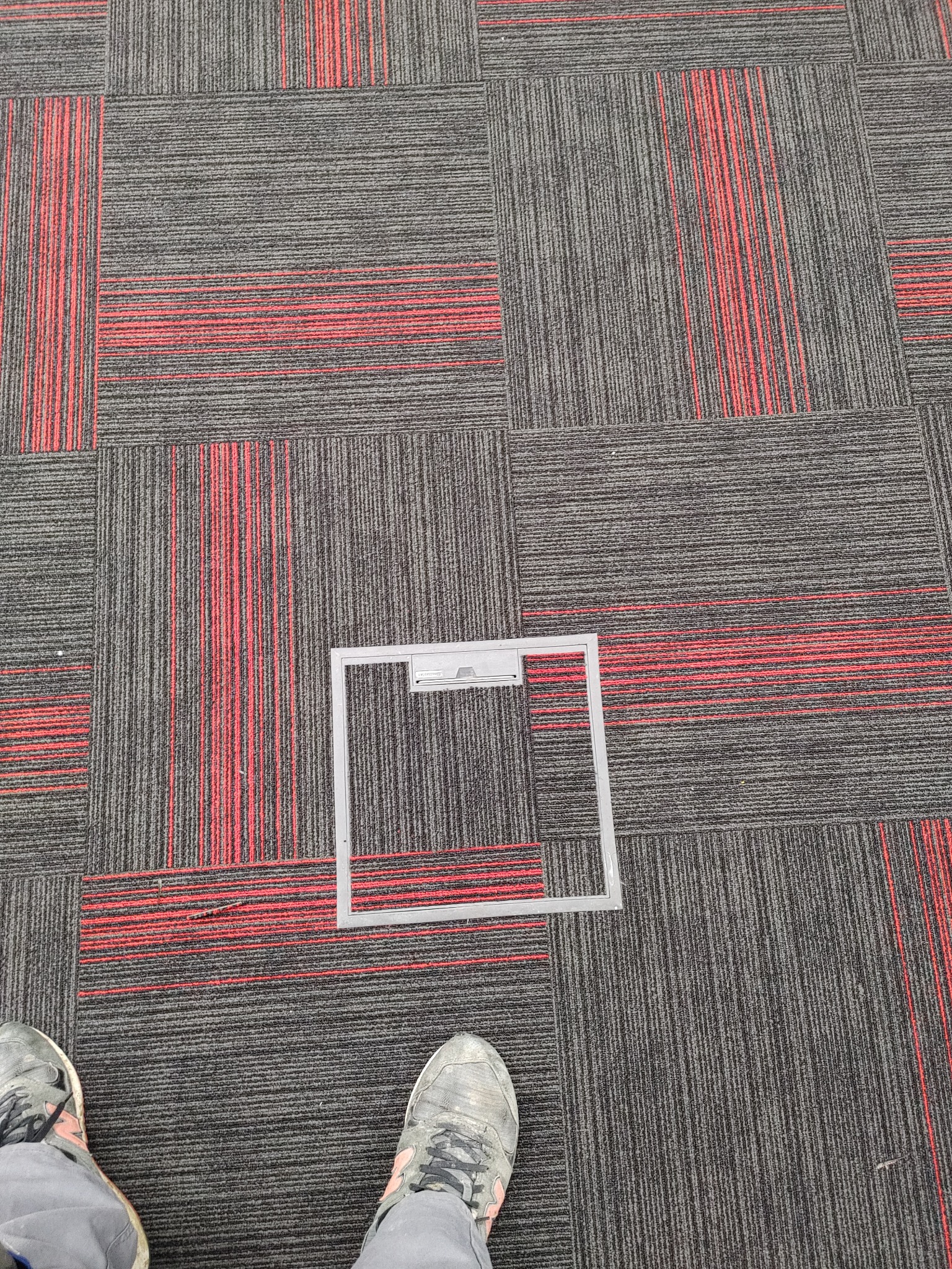 Szare płytki dywanowe z czerwonymi akcentami ułożone w kwadraty, z otwartą ramką dostępową do podłogi i fragmentem butów w dolnej części kadru.