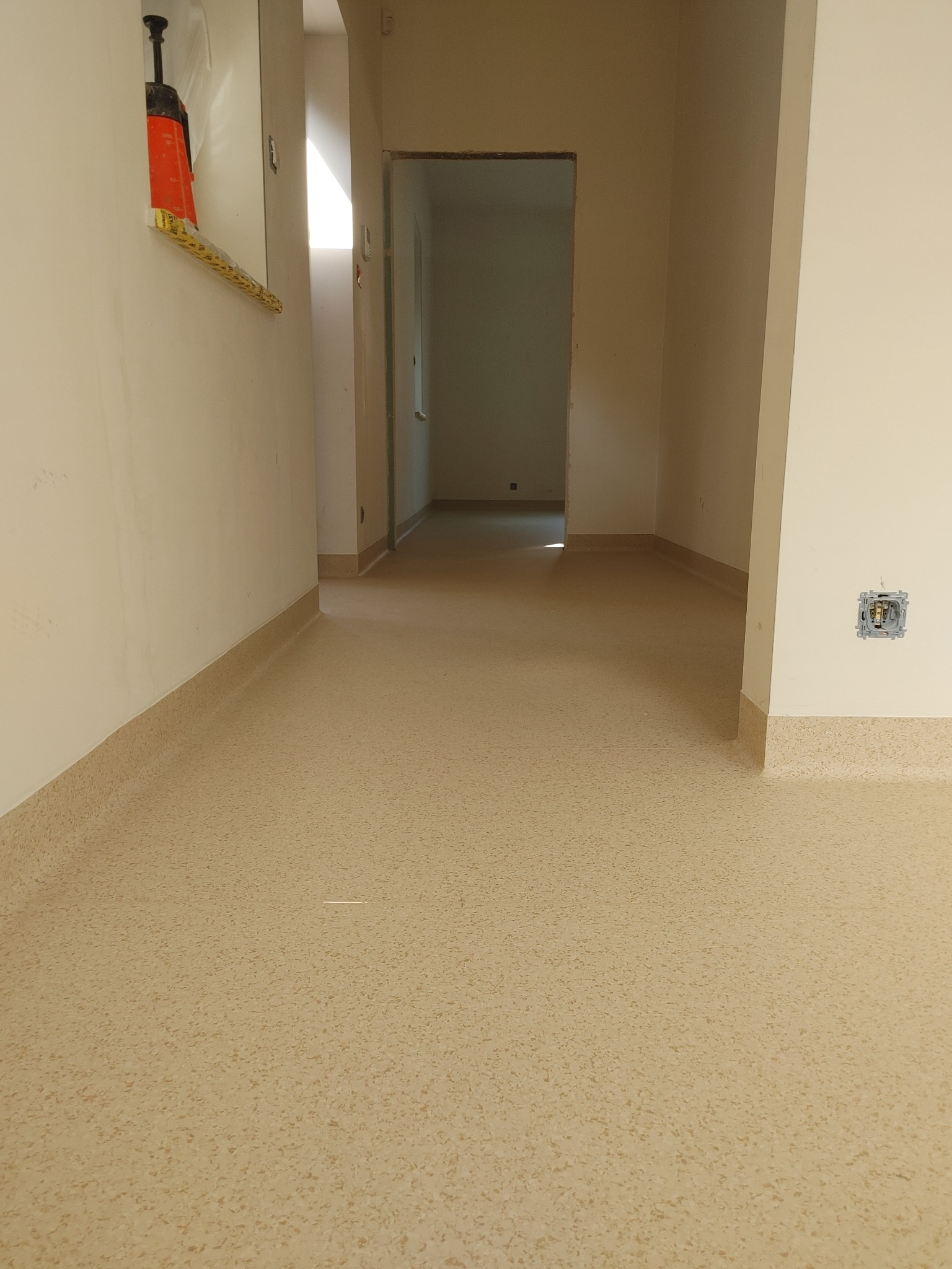 Beżowa wykładzina PCV ułożona w korytarzu, z widocznymi listwami przypodłogowymi i gniazdkiem elektrycznym bez osprzętu. W tle widoczne kolejne pomieszczenia.