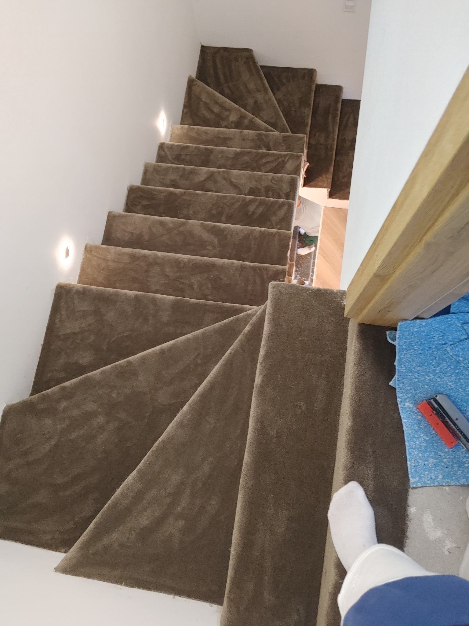 Kaskadowe schody wyłożone beżową wykładziną, widziane z góry. Fragment stopy w skarpetce na dole kadru. Oświetlenie punktowe na ścianie. Widoczne narzędzia pracy.