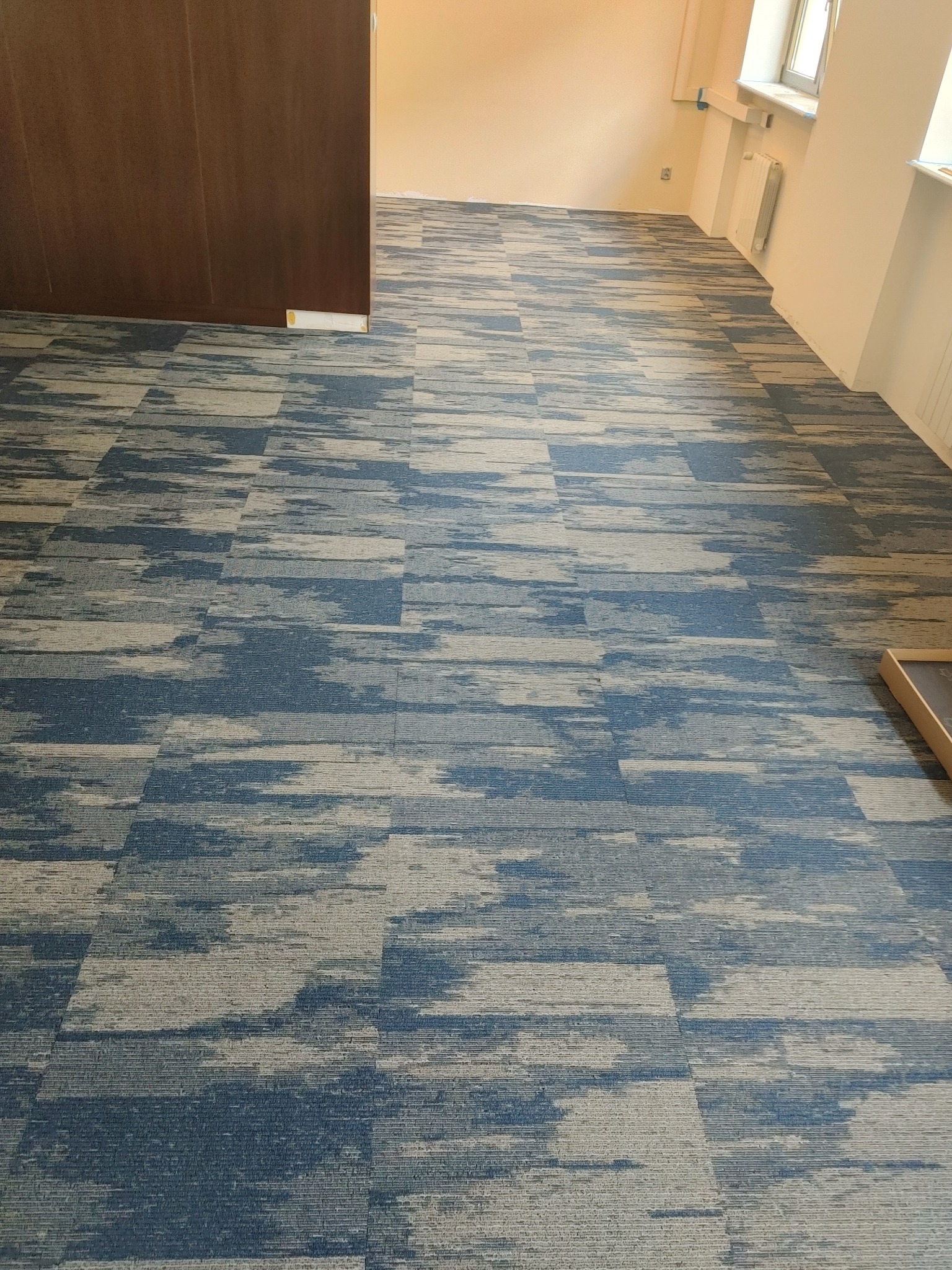 Wzorzysta, błękitno-szara wykładzina dywanowa w płytkach, ułożona w przestronnym wnętrzu z oknem i jasnymi ścianami. Widoczny fragment drzwi po lewej stronie.