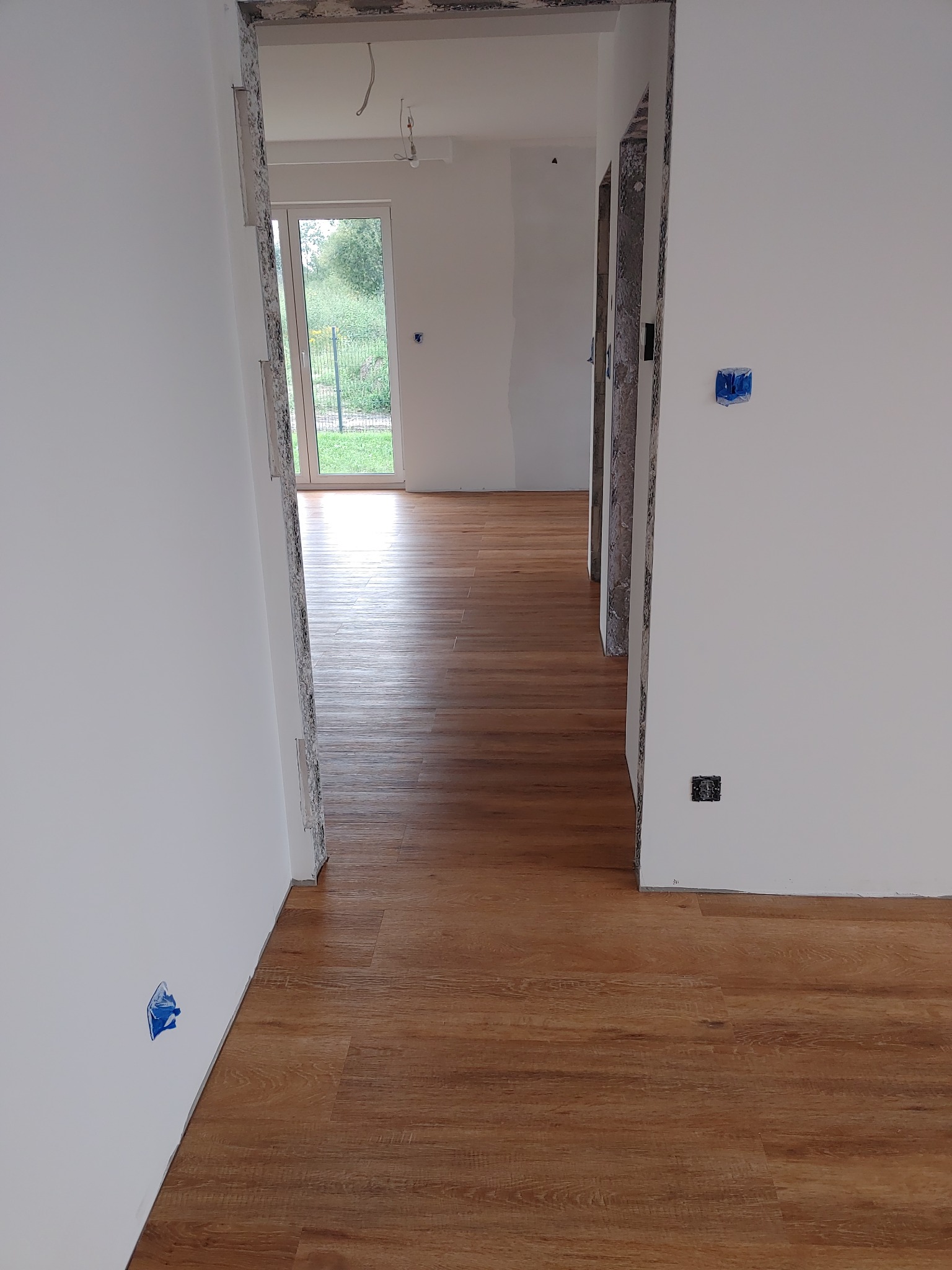 Ukończona podłoga z paneli w jasnym wnętrzu. Widoczne detale wykończenia i świeżo pomalowane ściany. Przejście do kolejnego pomieszczenia z widokiem na zieleń.