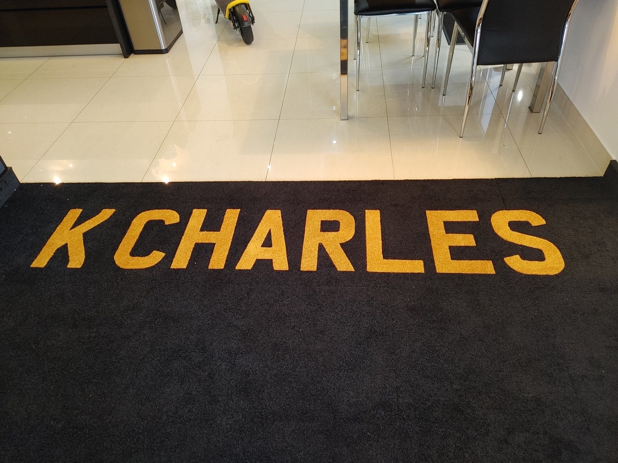 Czarna wykładzina z napisem 'K CHARLES' w złotym kolorze, leżąca na jasnych płytkach. W tle fragment skutera i krzesła.