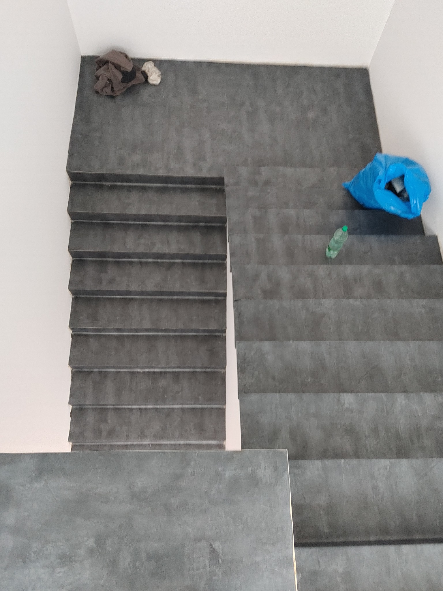 Szare schody wyłożone płytkami, widok z góry. Na schodach leżą śmieci: worek i butelka. Widoczny fragment ściany pomalowanej na biało.