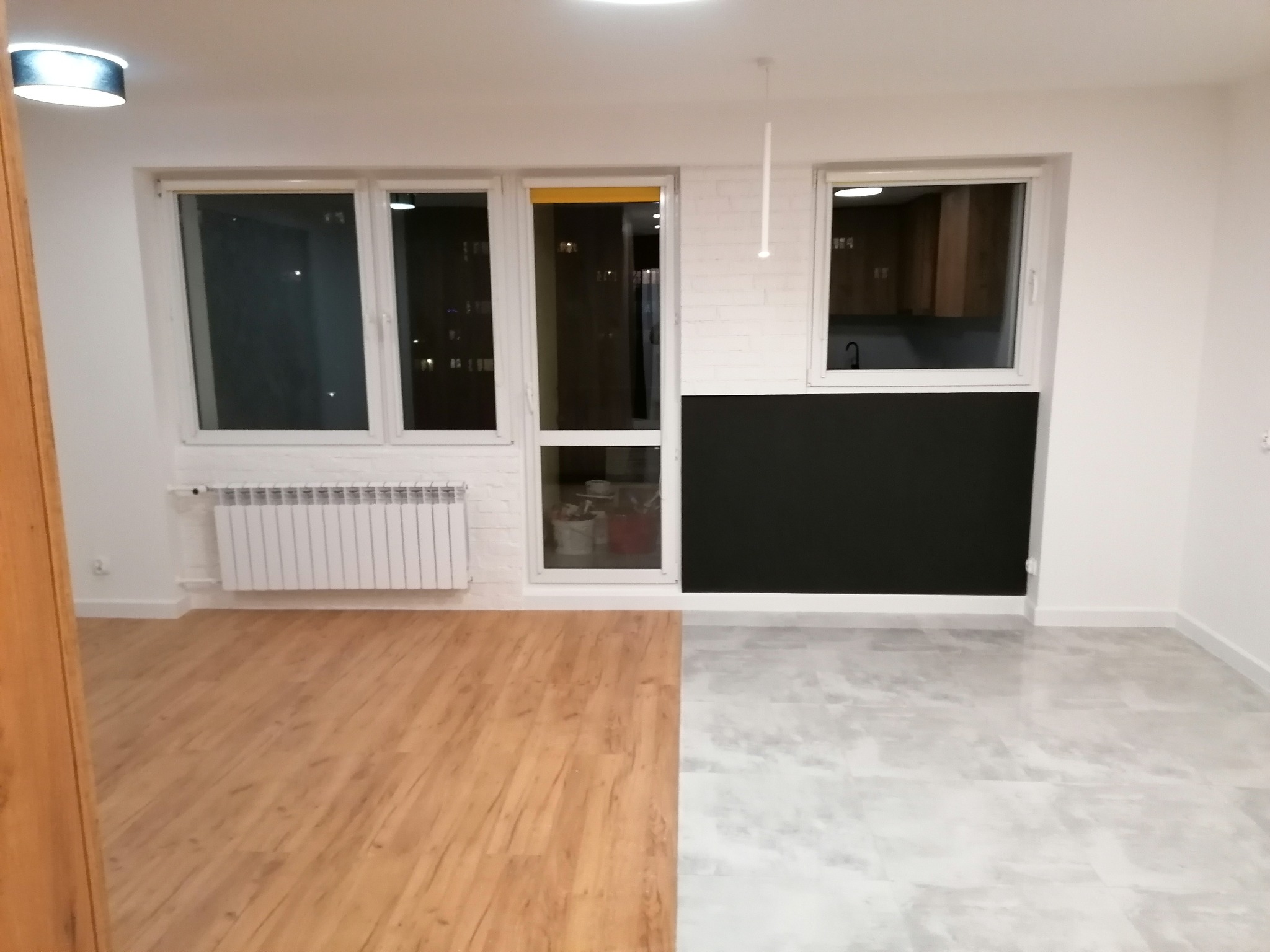 Pokój po remoncie z podłogą podzieloną na panele drewnopodobne i imitację betonu, białe ściany z czarnym pasem, grzejnik pod oknem.