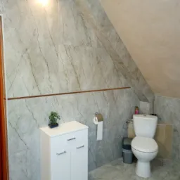 Wykończona łazienka na poddaszu ze skośnym sufitem, marmurowe płytki na ścianach i podłodze, biała szafka pod umywalkę z bonsai, toaleta z koszem na śmieci obok.