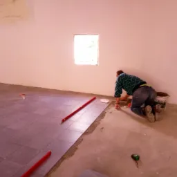 Układanie brązowych płytek podłogowych w pomieszczeniu z małym oknem, widoczne narzędzia i wiadro kleju Mozalit.