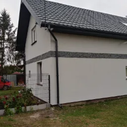 Elewacja domu jednorodzinnego z szarą, dekoracyjną listwą imitującą kamień, ciemną blachodachówką i orynnowaniem. W tle czerwony bus i zieleń ogrodu.