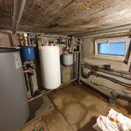 Piwnica z zainstalowanym systemem grzewczym: zbiornik akumulacyjny, biały zbiornik wyrównawczy, liczne rury z zaworami, okno z mleczną szybą. Widoczne narzędzia i szmatka na podłodze.