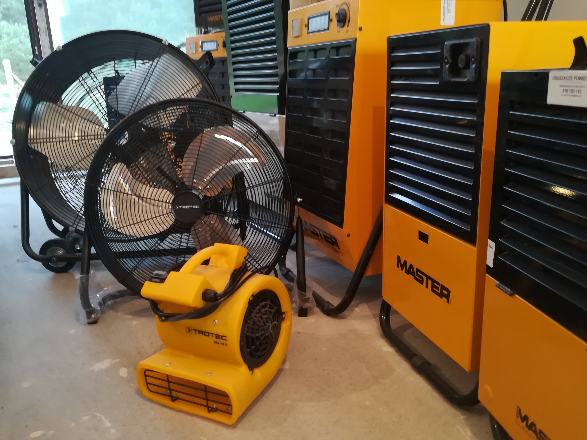 Żółte nagrzewnice i wentylatory budowlane różnych marek, w tym Trotec i Master, ustawione w pomieszczeniu z betonową podłogą. Widoczne detale konstrukcyjne i oznaczenia modeli.