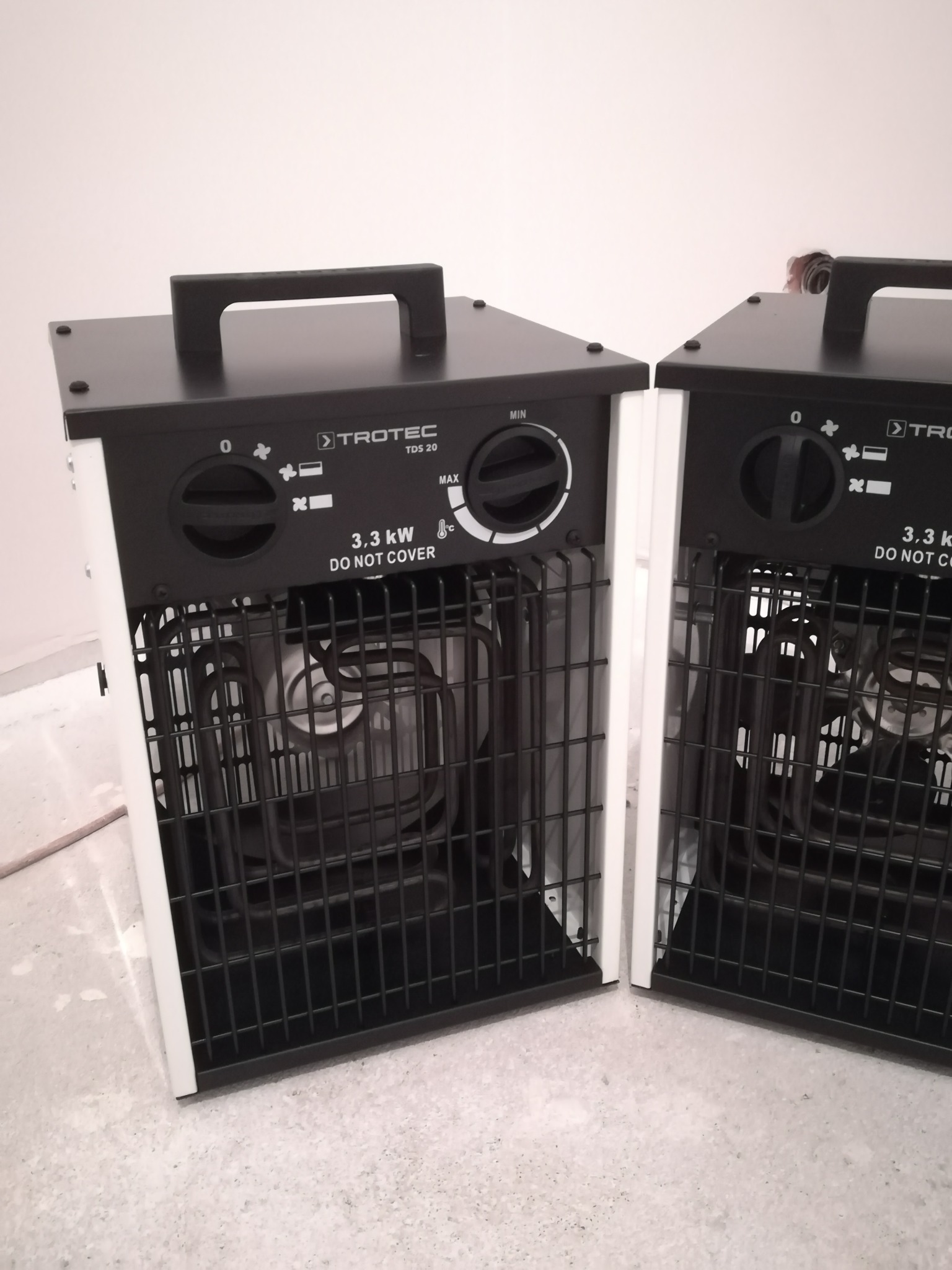 Dwa identyczne, przenośne grzejniki elektryczne Trotec TDS 20, z kratką ochronną i regulacją mocy, ustawione na betonowej posadzce w jasnym wnętrzu.