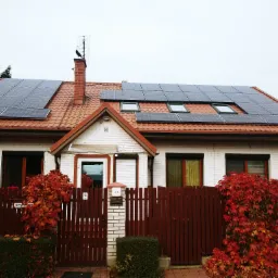 Biały dom z czerwonym dachem i panelami słonecznymi, widok z zewnątrz, jesienne liście na ogrodzeniu.