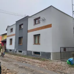 Dwukondygnacyjny budynek po remoncie elewacji, z szarą i białą częścią, drewnianą deską dekoracyjną, widoczne okna z roletami, chodnik z liśćmi i przechodząca osoba.