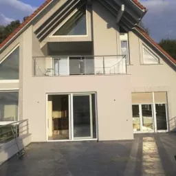 Dwupiętrowy dom z ocieploną elewacją w odcieniach beżu, z dużymi oknami i balkonem z metalową balustradą, widziany z poziomu tarasu wyłożonego kamienną płytą.