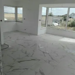 Nowo ułożona, błyszcząca podłoga z płytek imitujących marmur w jasnym pokoju z dużymi oknami wychodzącymi na pole i zabudowania.