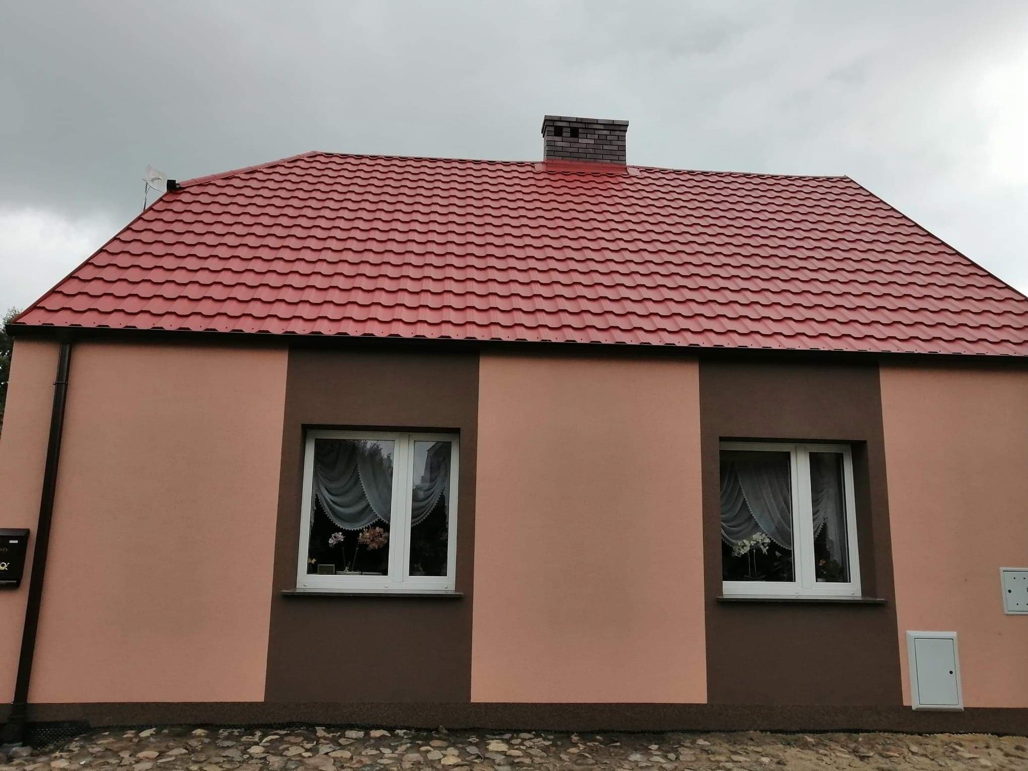 Dom jednorodzinny z nową, czerwoną blachodachówką i dwukolorową elewacją w odcieniach brązu i beżu, widok z poziomu gruntu, pochmurne niebo.
