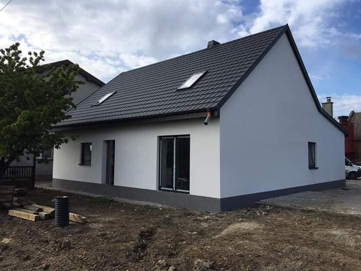 Wykończony dom jednorodzinny z ocieploną i pomalowaną na biało elewacją, ciemnoszarym dachem z oknami połaciowymi i szarą podmurówką, widoczny z zewnątrz na tle błękitnego nieba z chmurami.