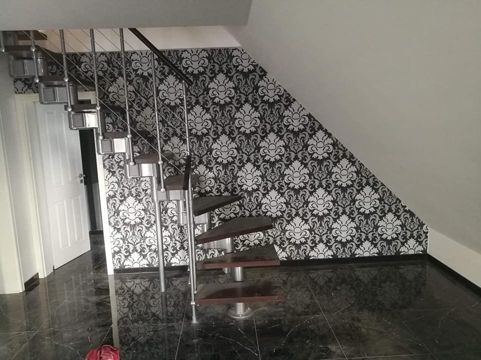 Spiralne schody z metalowymi poręczami i drewnianymi stopniami na tle ściany z czarno-białą tapetą w kwiatowe wzory, błyszcząca czarna podłoga.