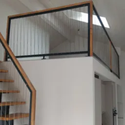 Nowoczesne schody z drewnianymi stopniami i balustradą z metalowych linek, prowadzące na antresolę z podobną balustradą, w jasnym wnętrzu z białymi ścianami i skośnym sufitem.