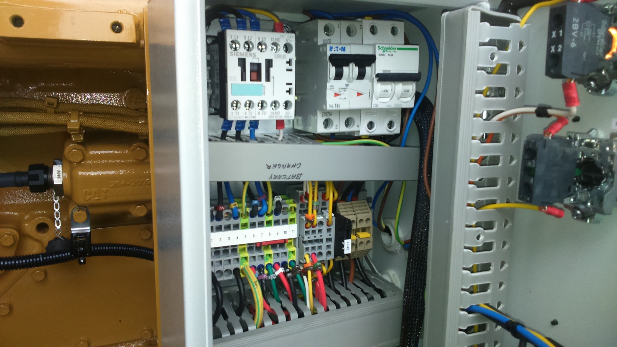 Otwarta szafa elektryczna z widocznymi elementami sterowniczymi Siemens i Schneider Electric, okablowaniem oraz fragmentem żółtej obudowy agregatu prądotwórczego CAT po lewej stronie.