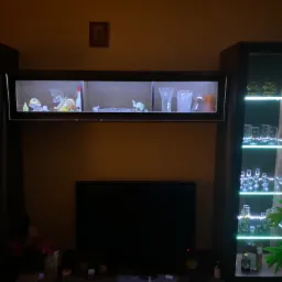 Ciemne wnętrze salonu z podświetlaną witryną i półką, na których ustawione są szklane naczynia i dekoracyjne figurki, a poniżej umieszczony jest telewizor.