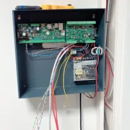 Otwarta skrzynka z elektroniką alarmową, widoczne płytki drukowane, zasilacz impulsowy, oraz wiązka kabli o różnych kolorach, w górnej części skrzynki leżą dwa śrubokręty