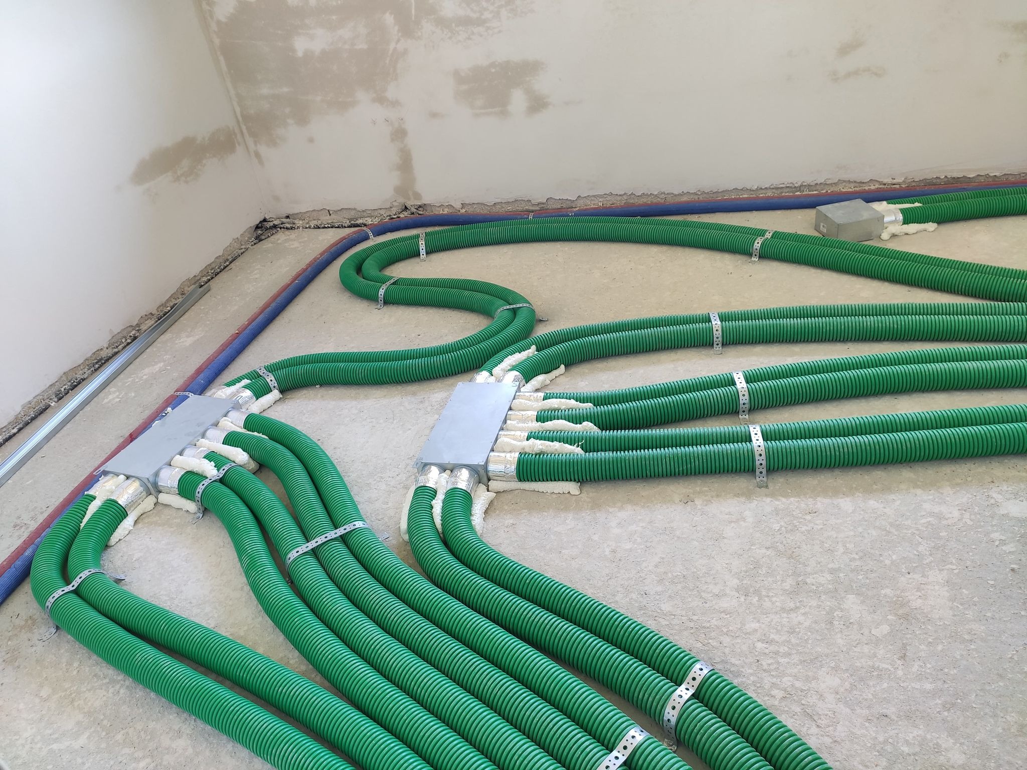 Rozprowadzenie zielonych, karbowanych rur wentylacyjnych z metalowymi obejmami w surowym wnętrzu budynku w trakcie budowy, widoczne również szare skrzynki rozdzielcze i fragment nieotynkowanej ściany.