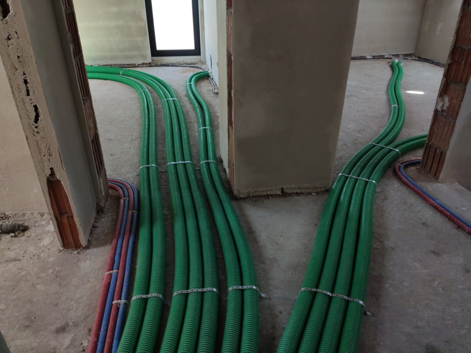 Zielone, karbowane rury wentylacyjne ułożone w wiązkach na surowym betonie w trakcie montażu w nowym budynku, zabezpieczone metalowymi opaskami, widoczne również rury instalacji wodnej.