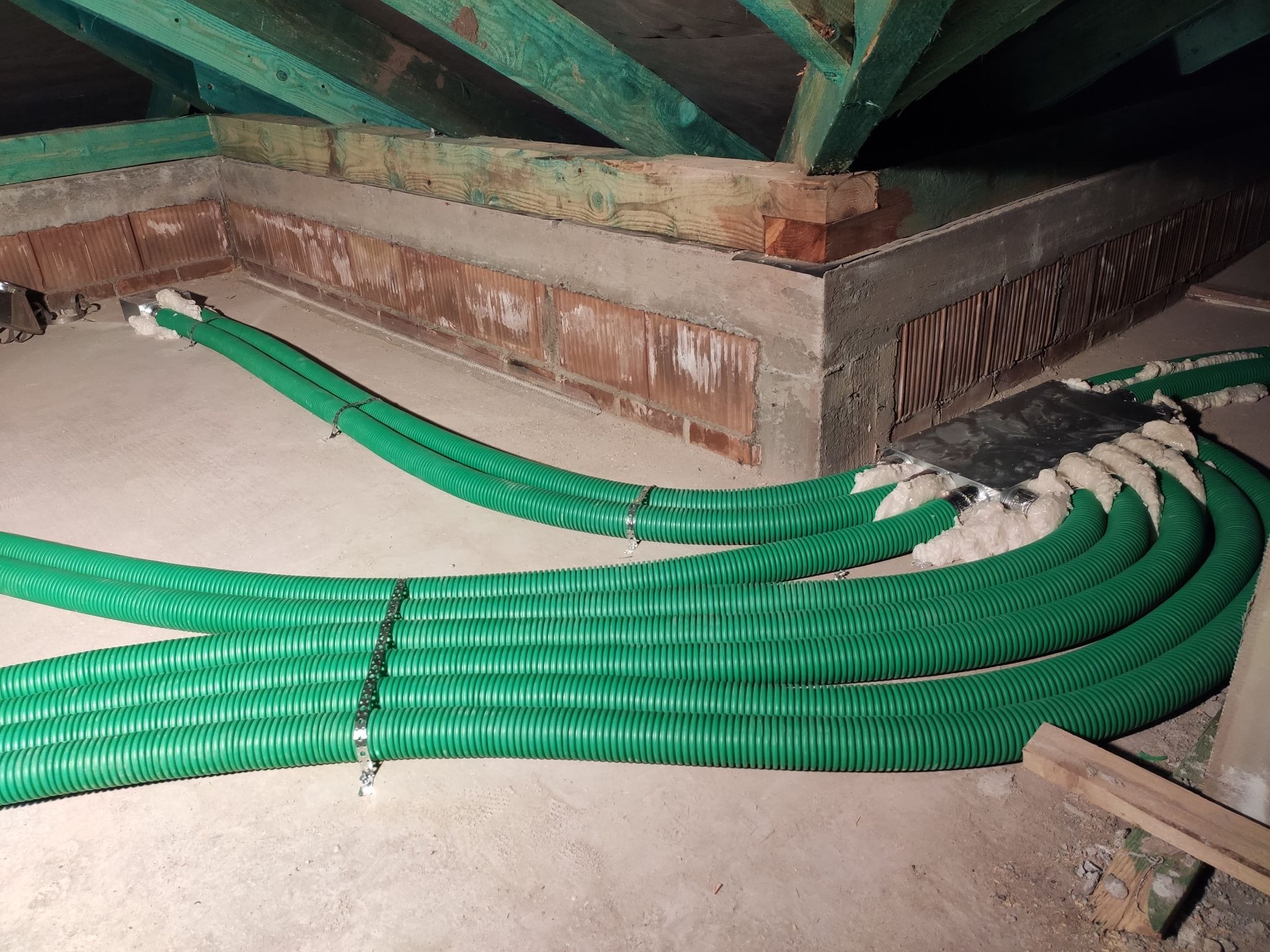 Zielone, karbowane rury wentylacyjne prowadzone w przestrzeni poddasza, zabezpieczone pianką montażową w miejscach połączeń, widoczne drewniane elementy konstrukcji dachu.