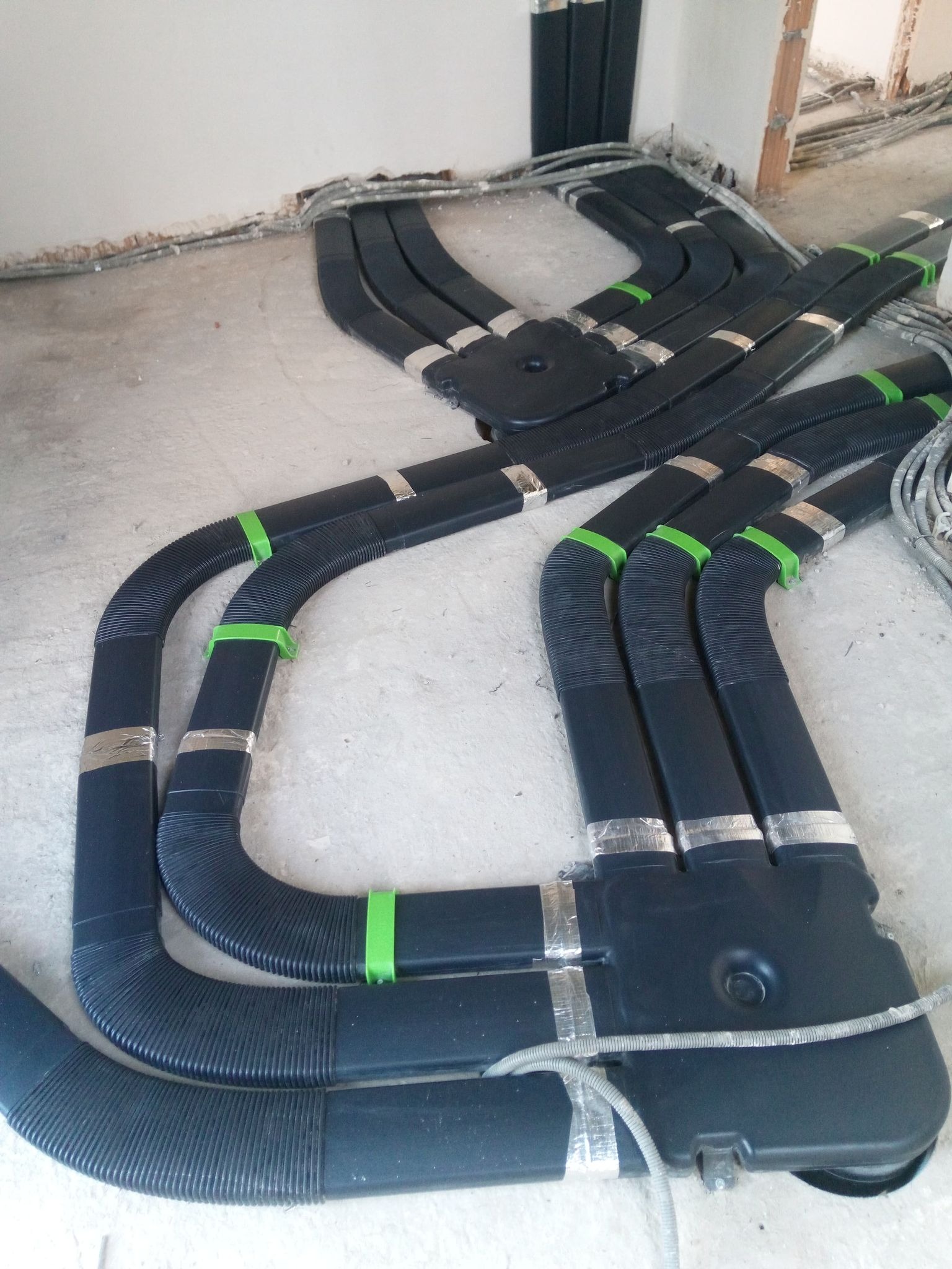 Czarna, elastyczna instalacja przewodów wentylacyjnych z zielonymi i srebrnymi taśmami mocującymi, ułożona na surowym betonie w trakcie montażu w budynku.