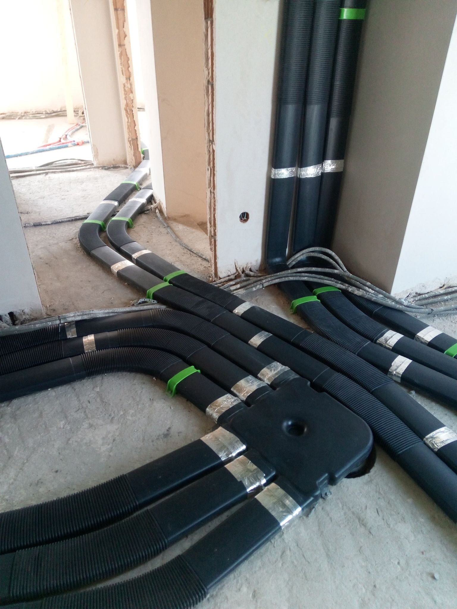 Czarny system rur wentylacyjnych z zielonymi i srebrnymi elementami mocującymi, ułożony na surowym betonowym podłożu w trakcie instalacji w budynku.