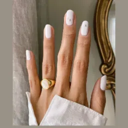 Dłoń z białym manicurem z subtelnymi złotymi zdobieniami w kształcie gwiazdek, ozdobiona złotym pierścieniem z perłowym oczkiem, na tle beżowej zasłony i fragmentu złotej ramy lustra.