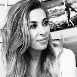 Portret kobiety z długimi, pofalowanymi włosami w odcieniach blondu, z delikatnym makijażem oczu i ust. Zdjęcie czarno-białe, z ostrością na twarz i włosy. W tle widoczny obraz z motywem morskim.