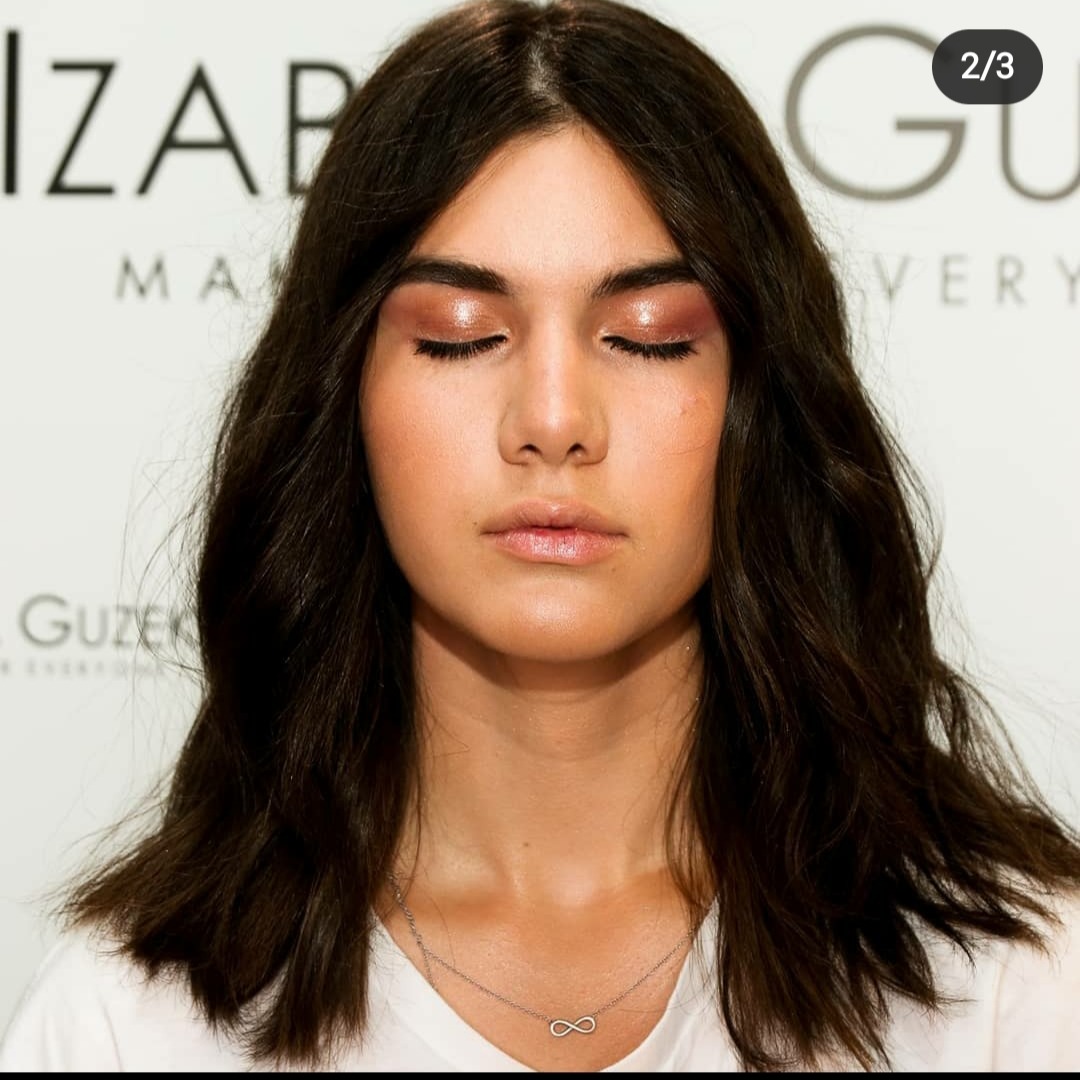 Portret kobiety z zamkniętymi oczami, prezentujący delikatny, błyszczący cień do powiek w odcieniach brązu i różu, z subtelnie podkreślonymi ustami i widocznym srebrnym naszyjnikiem z symbolem...