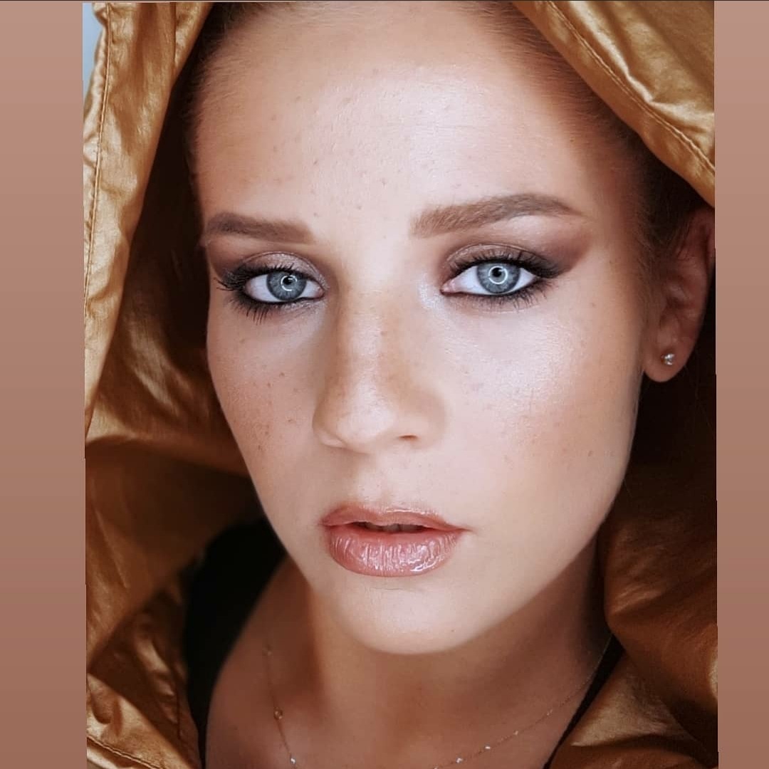 Portret kobiety z intensywnym makijażem oczu w odcieniach brązu i błyszczącymi ustami, ubrana w złotą kurtkę z kapturem, podkreślający jej niebieskie oczy.